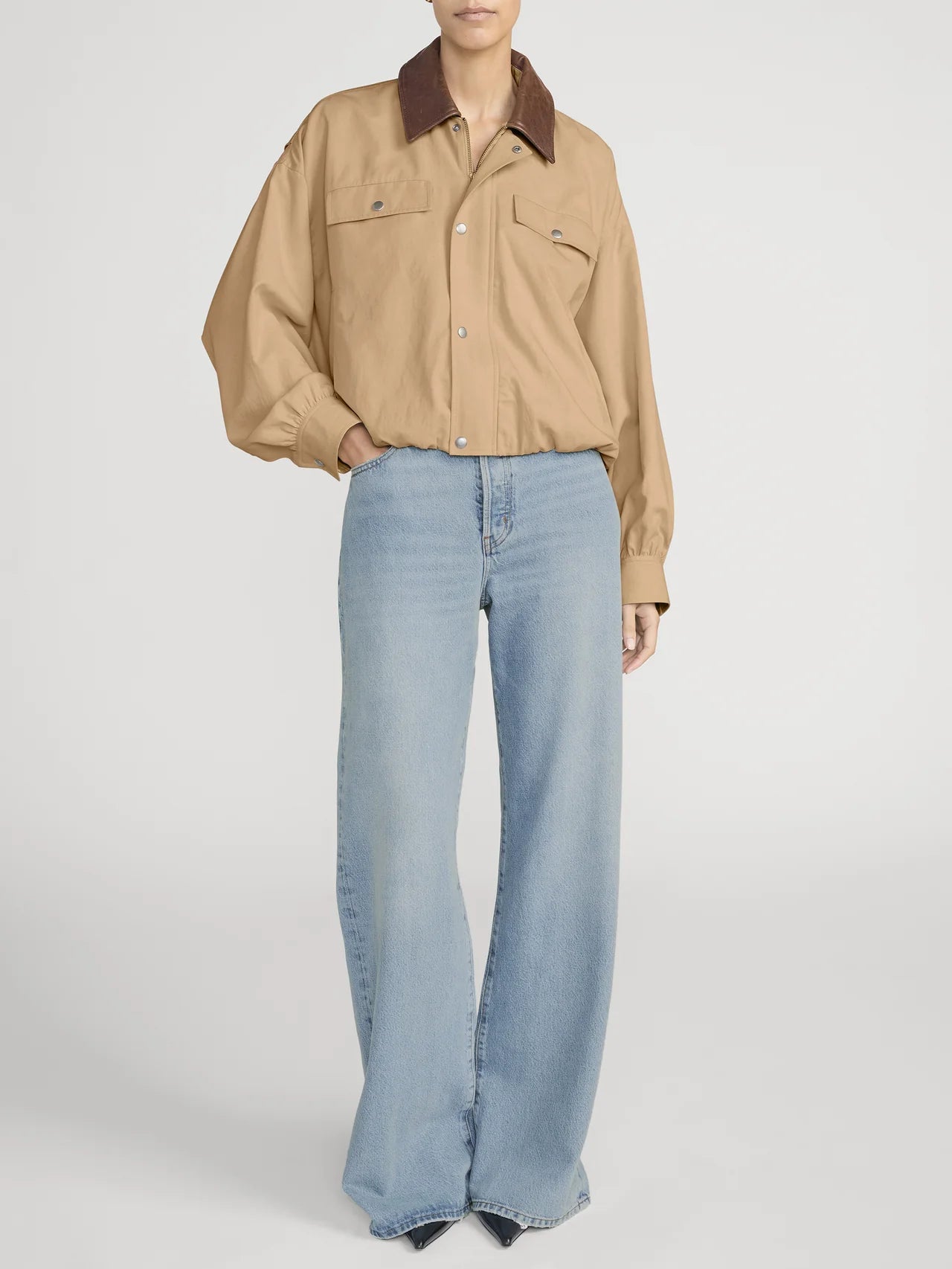 Person wearing a beige jacket with a brown collar and light blue jeans on a white background