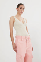 A female model wearing a beige contour knitted tank top with deep neckline and thin straps, paired with pink trousers with pockets.