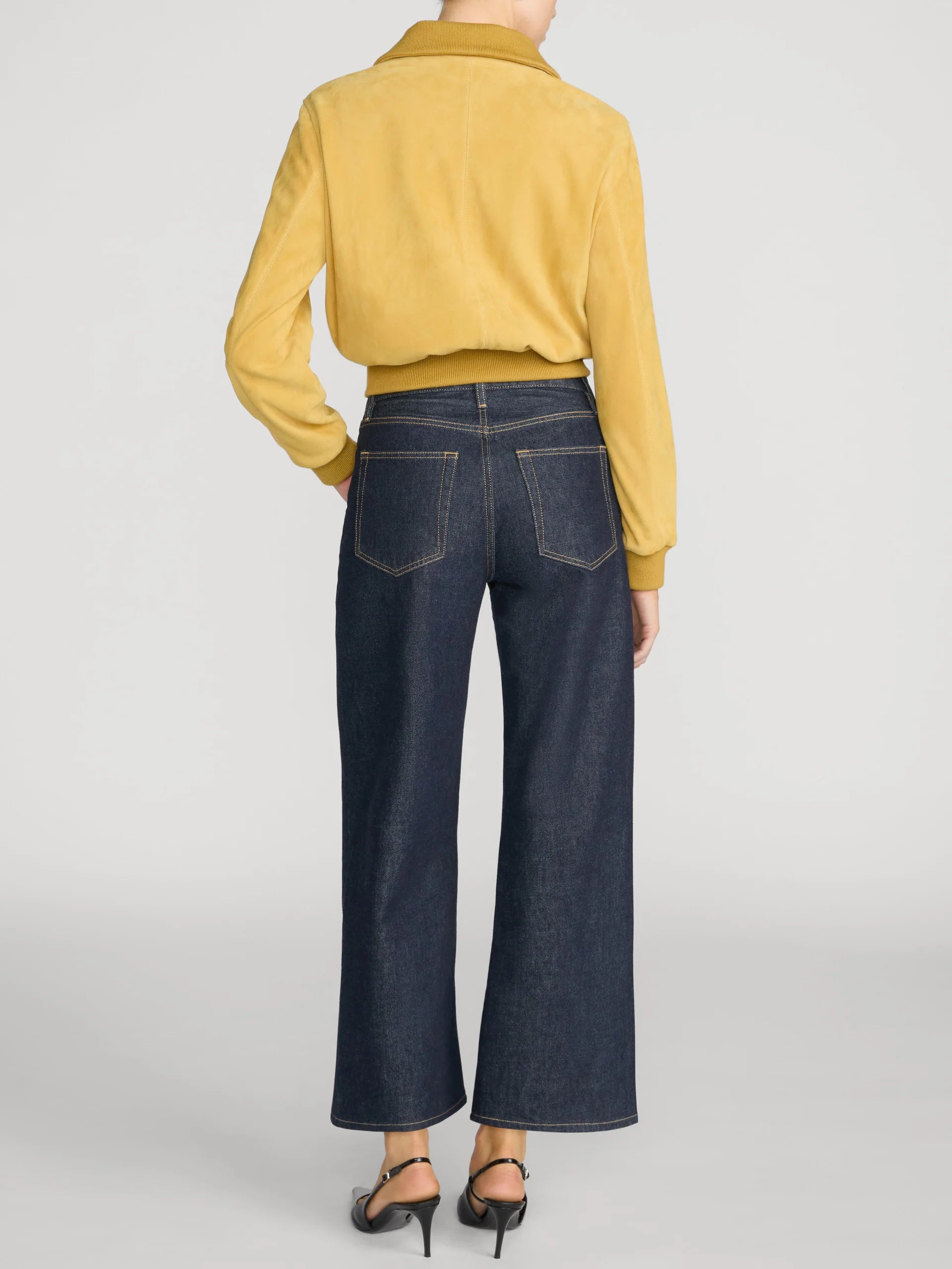 Rear view of a model wearing dark blue wide-leg jeans, a mustard yellow cropped knit top, and black high-heeled sandals against a light gray studio background.