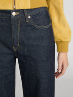 Close-up of a person wearing dark indigo denim jeans with contrast yellow stitching, paired with a mustard yellow jacket, against a light gray background.