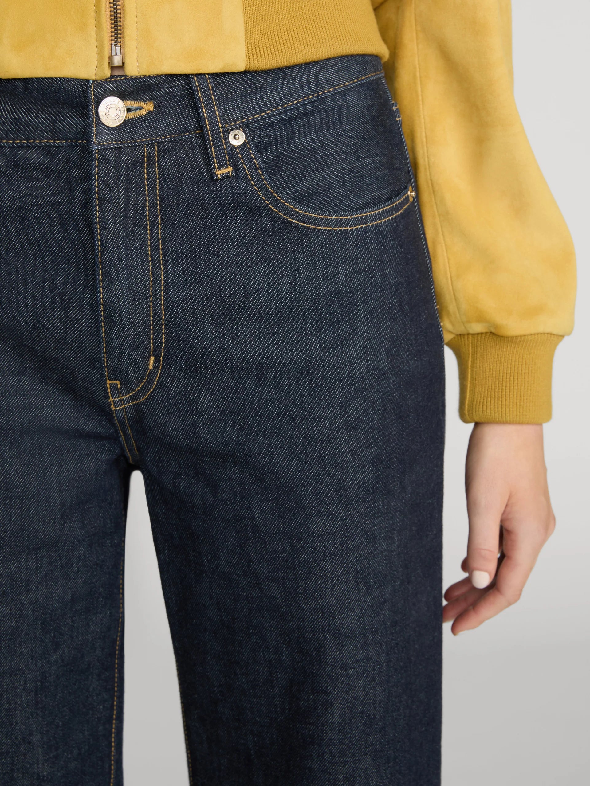 Close-up of a person wearing dark indigo denim jeans with contrast yellow stitching, paired with a mustard yellow jacket, against a light gray background.