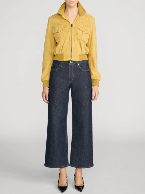A model wearing a cropped mustard yellow zip-up jacket with flap chest pockets and dark blue wide-leg cropped jeans, paired with black pointed heels, against a light gray studio background.