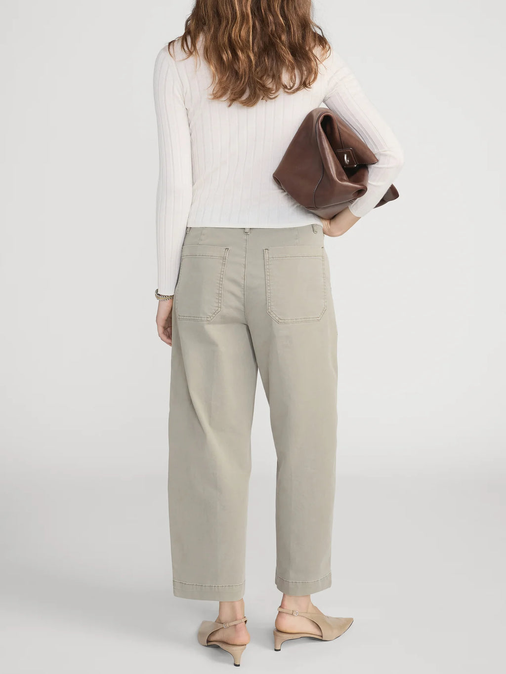 Woman wearing beige pants and a white top, holding a brown clutch against a light background