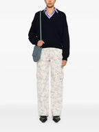 A model wearing a black knitted sweater with a V-neckline and ribbed trims, holding a blue bag and standing against a plain background.