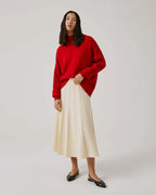 A woman wearing a red sweater and a beige flared midi skirt with ribbed panels, standing against a plain background.