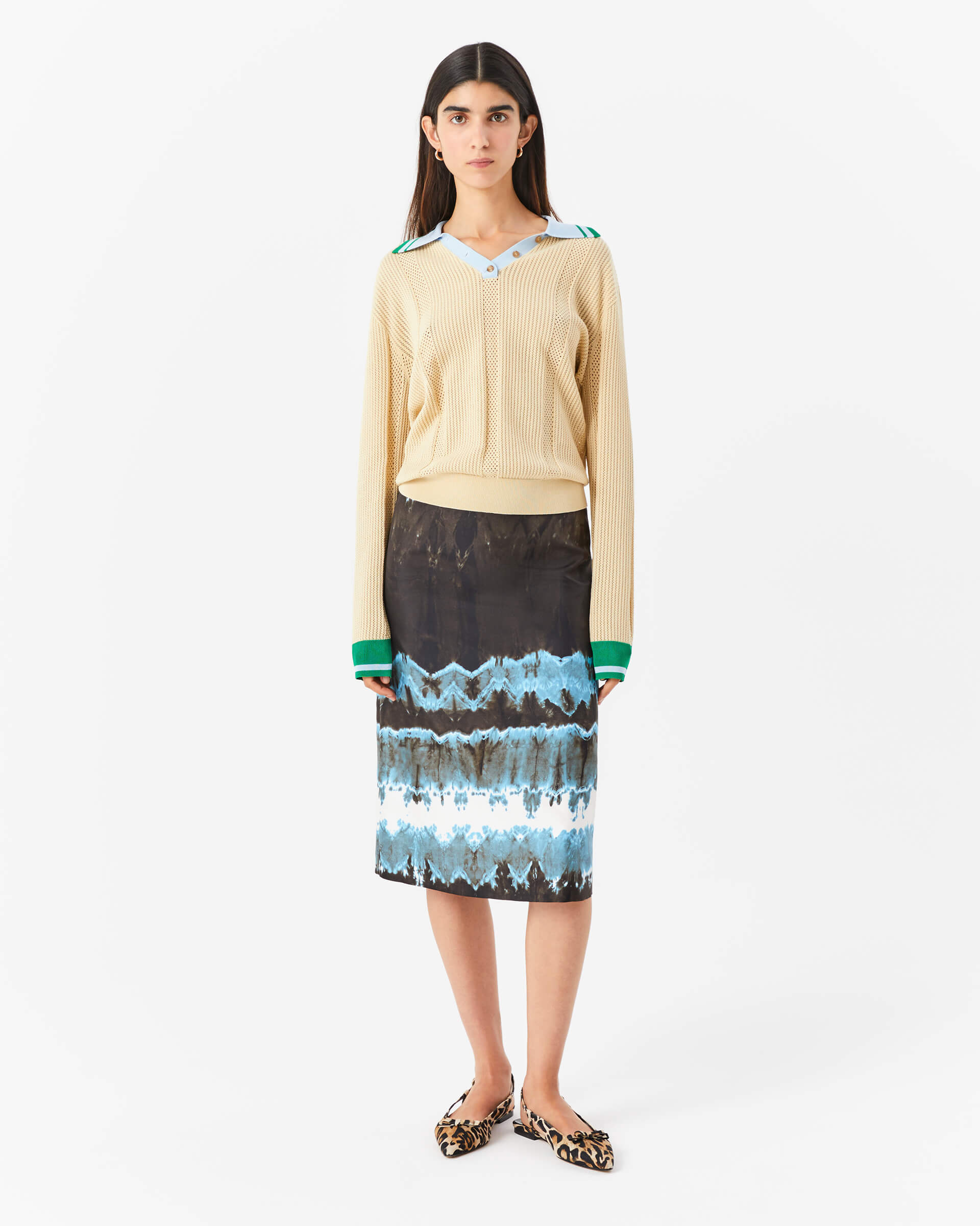 A woman standing wearing a beige jacket over a blue and green printed silk twill skirt with a black belt.