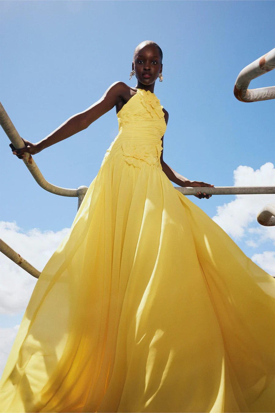 Woman in a yellow dress standing on metal bars against a blue sky.