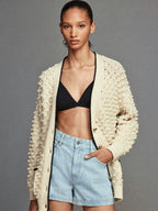 A woman wearing a beige cardigan with a crew neckline and long sleeves, paired with a black top and blue denim shorts.