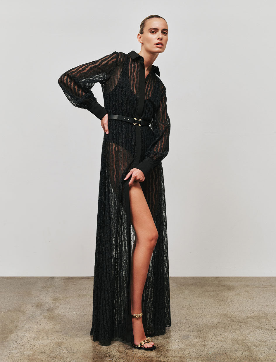Woman wearing a Rebecca vallance   seraphina maxi black sheer dress with a belt and slit on a plain background