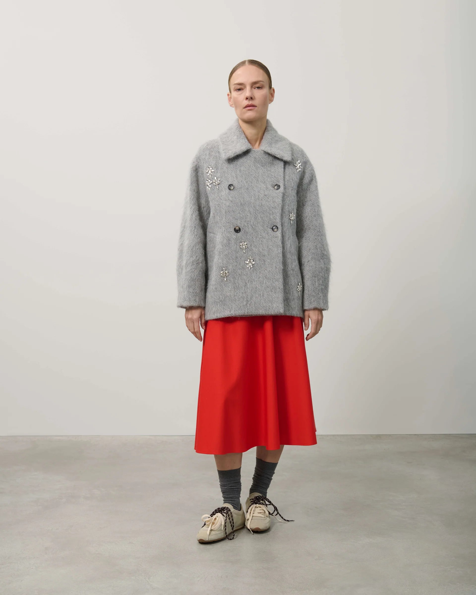 Person wearing a gray coat with floral embroidery and a red skirt on a plain background