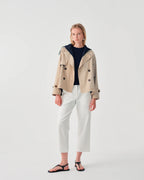 Woman wearing a beige trench coat, navy sweater, white pants, and black sandals on a light gray background
