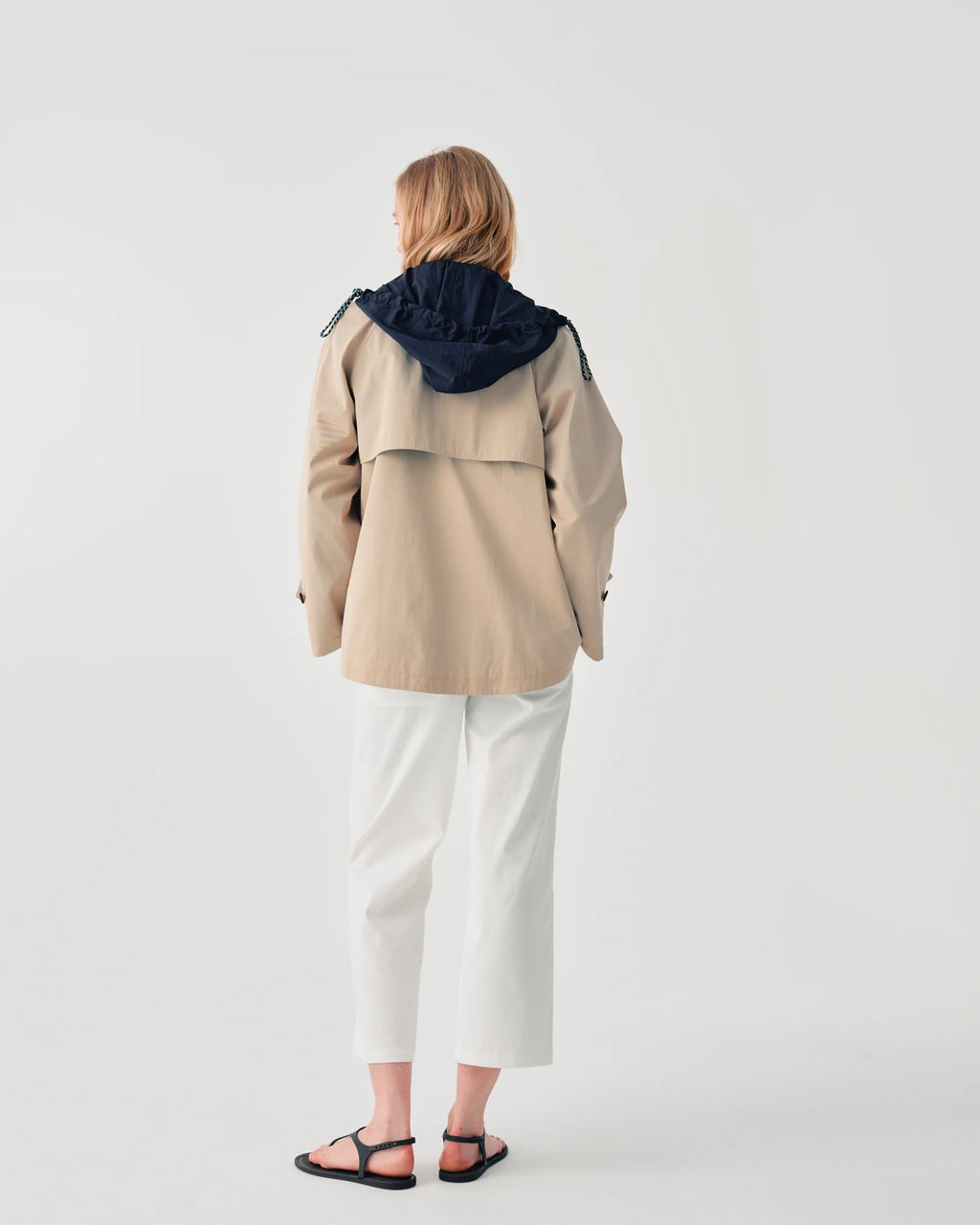 A woman viewed from behind wearing a beige hooded jacket with a contrasting dark navy hood, white cropped pants, and black sandals, set against a plain light gray studio background.