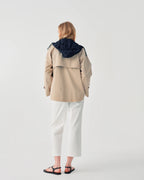 A woman viewed from behind wearing a beige hooded jacket with a contrasting dark navy hood, white cropped pants, and black sandals, set against a plain light gray studio background.