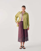 A model stands against a light gray studio background wearing a light olive green double-breasted jacket over a pale pink top, paired with a dusty purple wide-leg skirt featuring a patterned hem and black sandals.