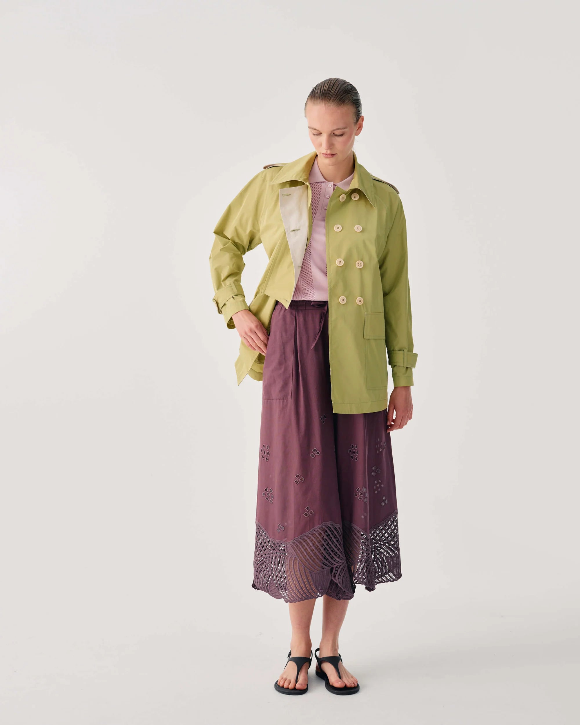 A model stands against a light gray studio background wearing a light olive green double-breasted jacket over a pale pink top, paired with a dusty purple wide-leg skirt featuring a patterned hem and black sandals.