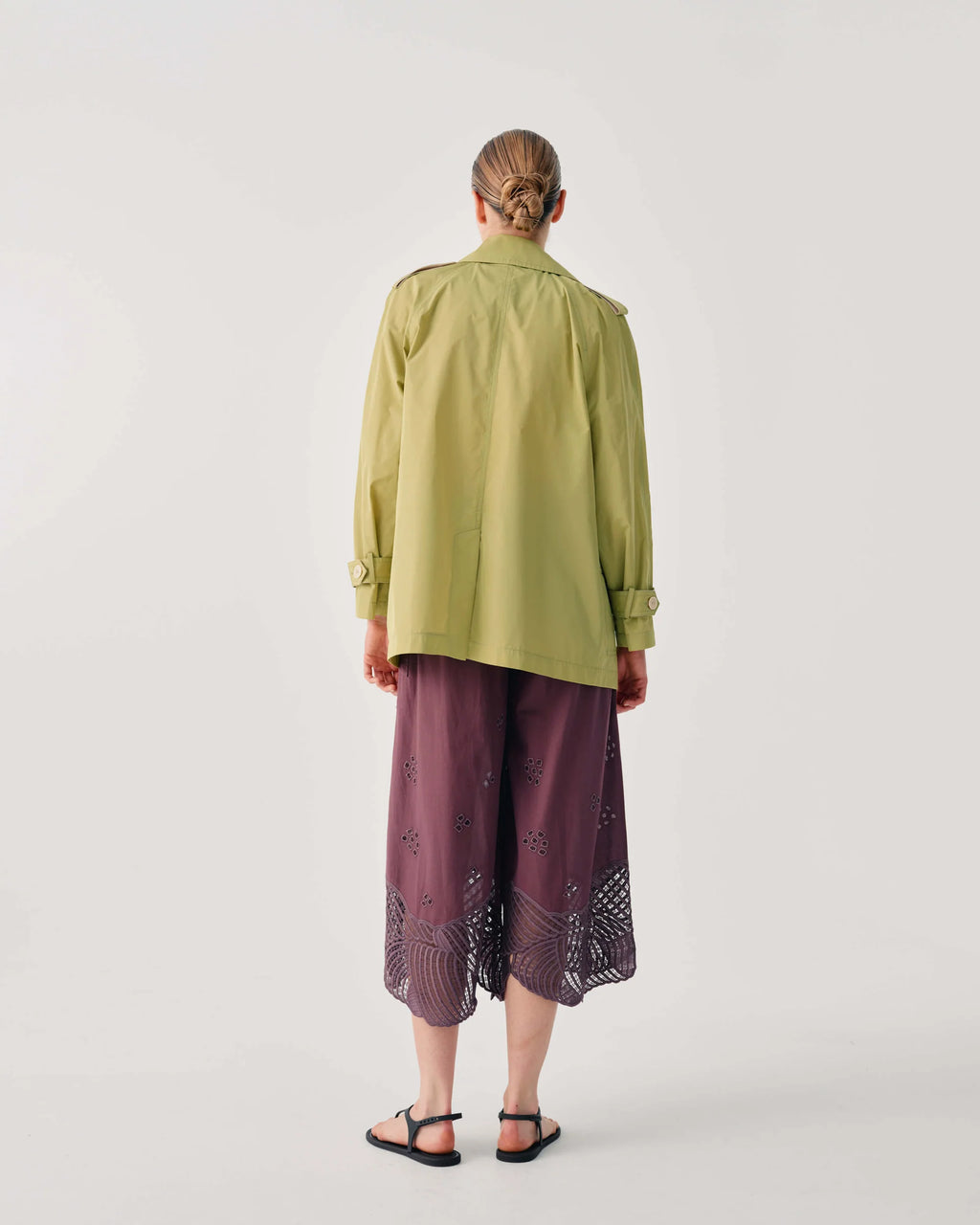 A model viewed from the back wearing a light olive green long-sleeve button-up shirt and dark purple wide-leg cropped pants with lace trim, paired with black sandals, against a light gray studio background.