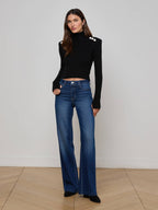 Woman wearing a black turtleneck and blue jeans standing against a plain white wall.