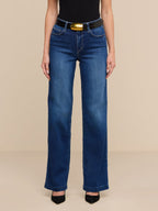 A model wearing a pair of dark blue wide-leg jeans with a black belt featuring a prominent gold buckle, styled with a black top and black heels against a solid beige background.