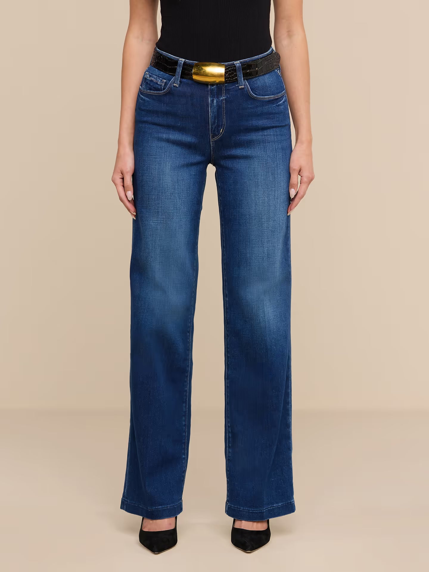 A model wearing a pair of dark blue wide-leg jeans with a black belt featuring a prominent gold buckle, styled with a black top and black heels against a solid beige background.
