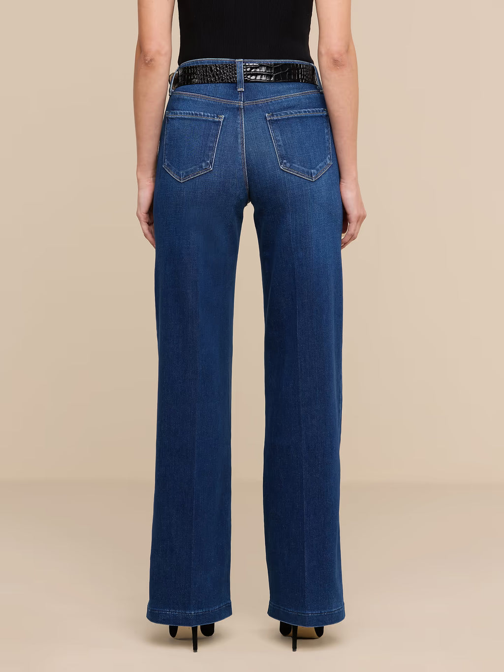 A model viewed from the back wearing dark blue wide-leg jeans with a black belt, a black bodysuit, and black heels, set against a solid beige studio background.