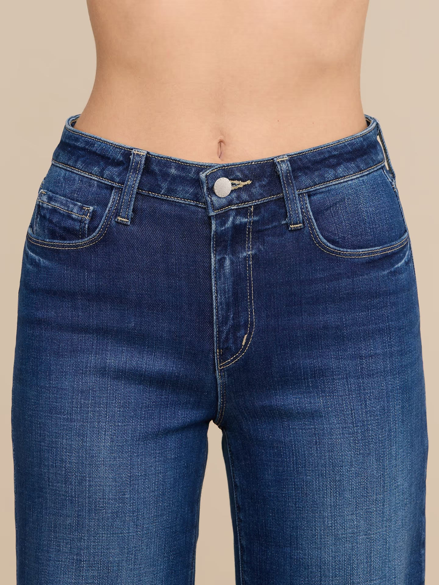 Close-up of a person wearing dark blue denim jeans, showing the waist, hips, and front pockets against a plain beige studio background.