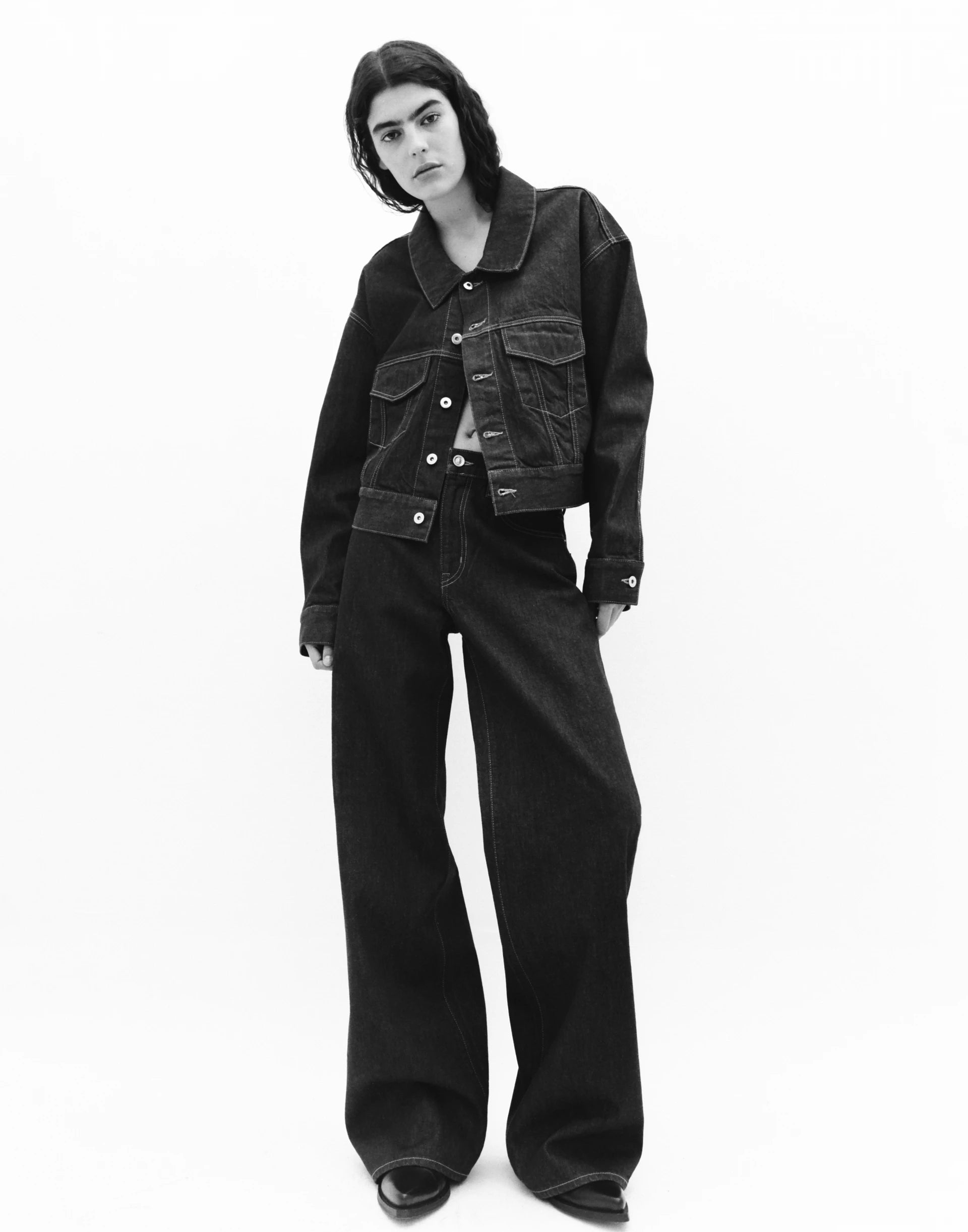 A person standing against a white background, wearing a black Selena wide-leg jeans and a matching jacket with a mid-rise waist and long sleeves.