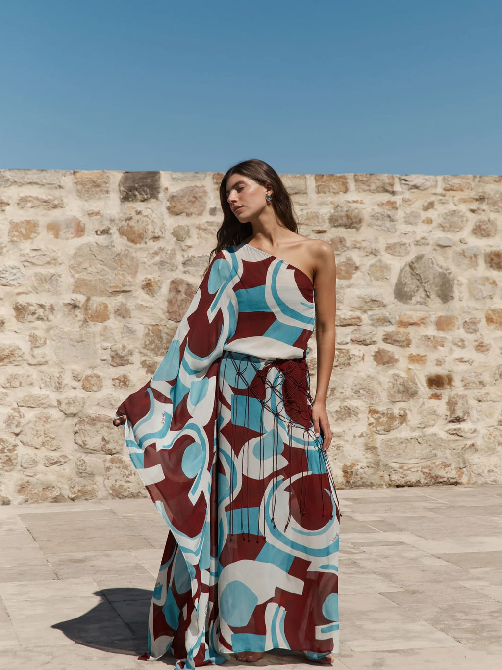 A woman stands outdoors against a stone wall under a clear blue sky, wearing a one-shoulder maxi dress with a bold abstract geometric print in teal, burgundy, cream, and white.