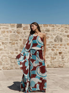 A woman stands outdoors against a stone wall under a clear blue sky, wearing a one-shoulder maxi dress with a bold abstract geometric print in teal, burgundy, cream, and white.