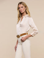 Woman wearing a white blouse and pants with a brown belt against a beige background