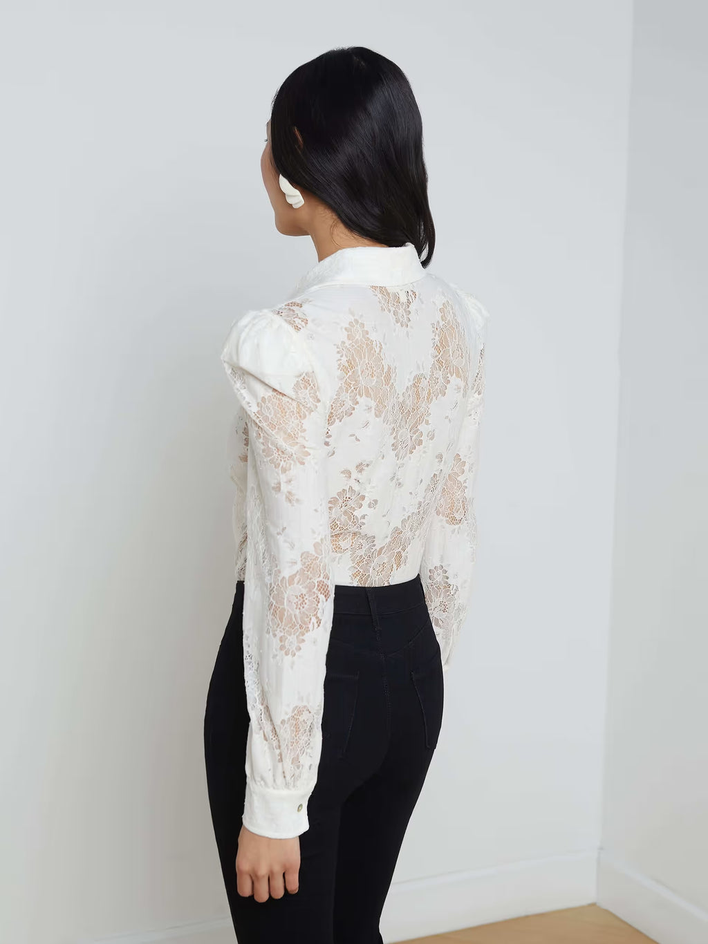 Person wearing a white lace blouse and black pants against a plain white wall.