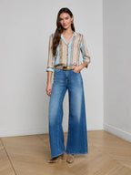 Woman wearing a light blue blouse and blue jeans standing in a room with wooden flooring.