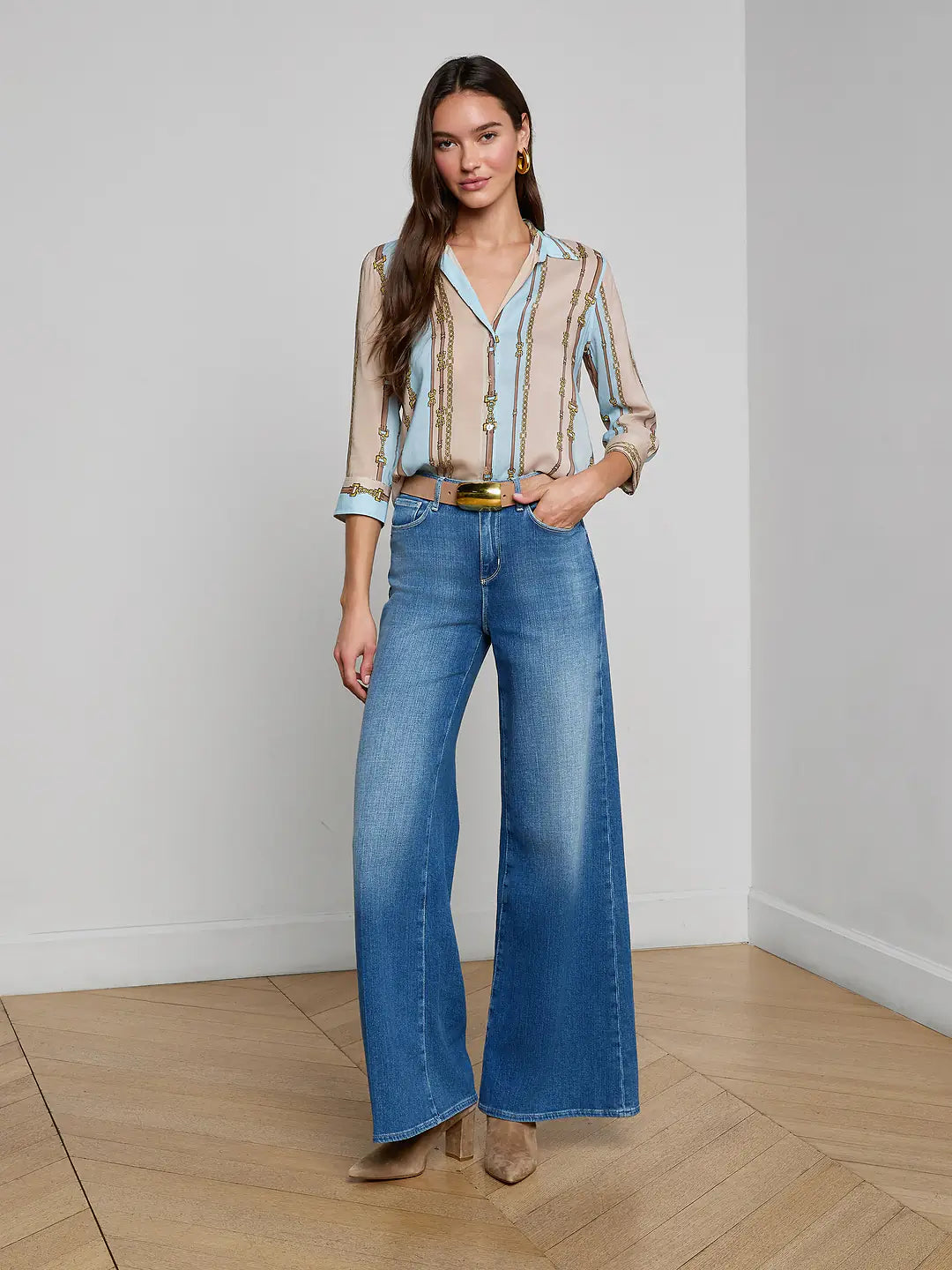 Woman wearing a light blue blouse and blue jeans standing in a room with wooden flooring.