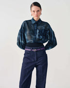 A model wearing a dark blue patterned sheer long-sleeve blouse tucked into dark blue trousers, accessorized with a purple belt, against a plain light gray background.
