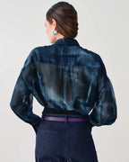 A woman shown from the back wearing a sheer dark blue patterned blouse with long sleeves, tucked into dark navy trousers with a purple belt.