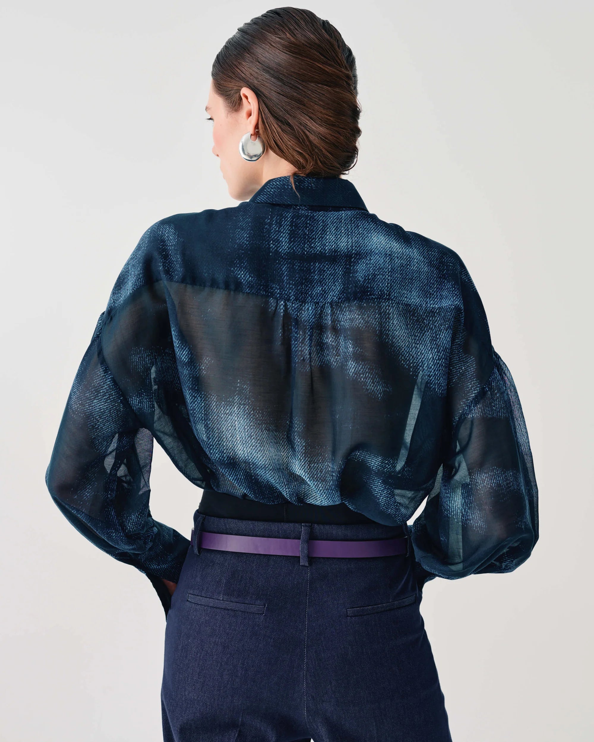 A woman shown from the back wearing a sheer dark blue patterned blouse with long sleeves, tucked into dark navy trousers with a purple belt.