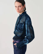A model wearing a dark blue sheer long-sleeve blouse with ruffle details and a textured pattern, paired with dark blue jeans and a purple belt, against a plain light gray background.