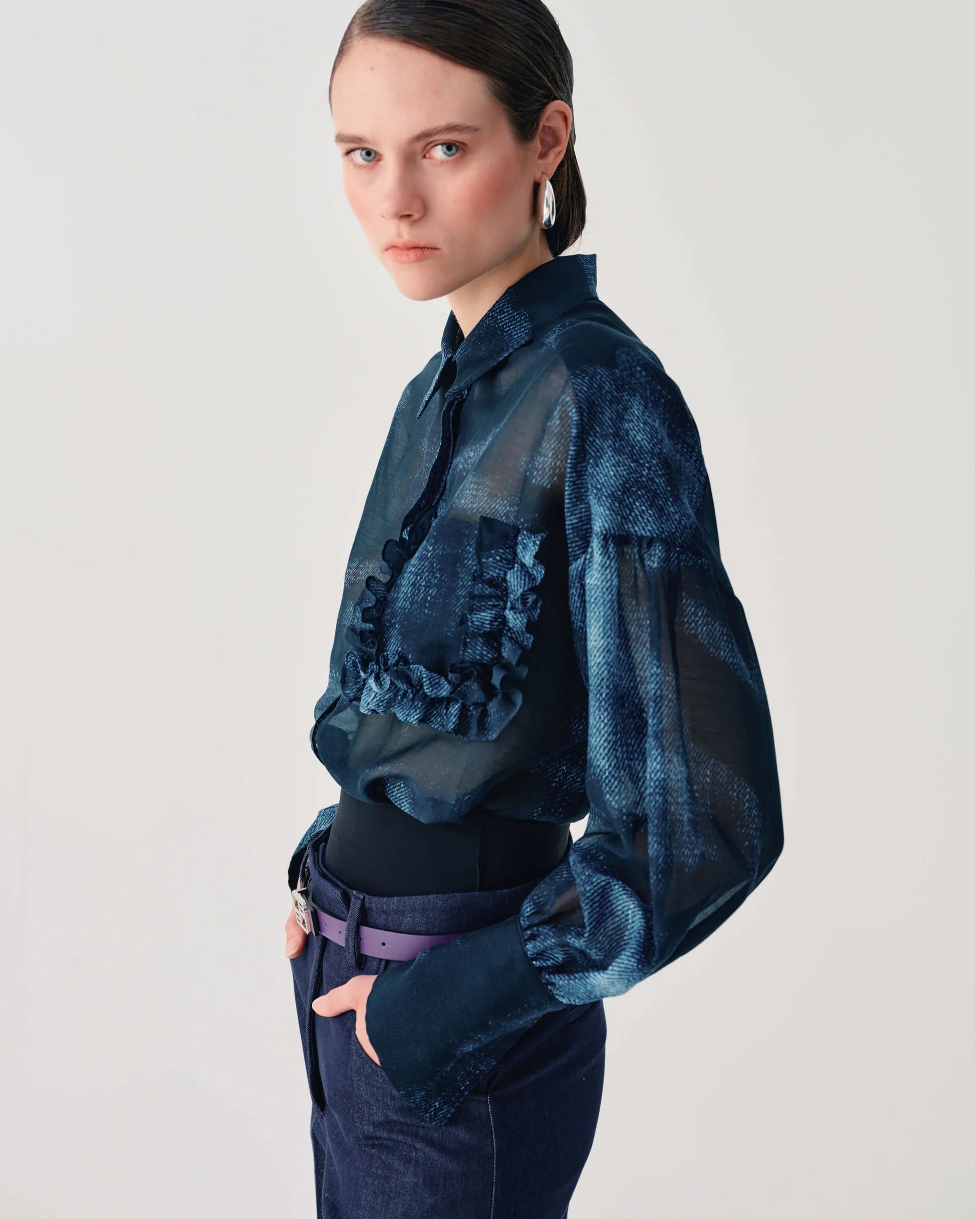 A model wearing a dark blue sheer long-sleeve blouse with ruffle details and a textured pattern, paired with dark blue jeans and a purple belt, against a plain light gray background.
