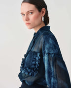 A model wearing a dark blue textured sheer blouse with ruffle details on the front, photographed against a plain light gray background.