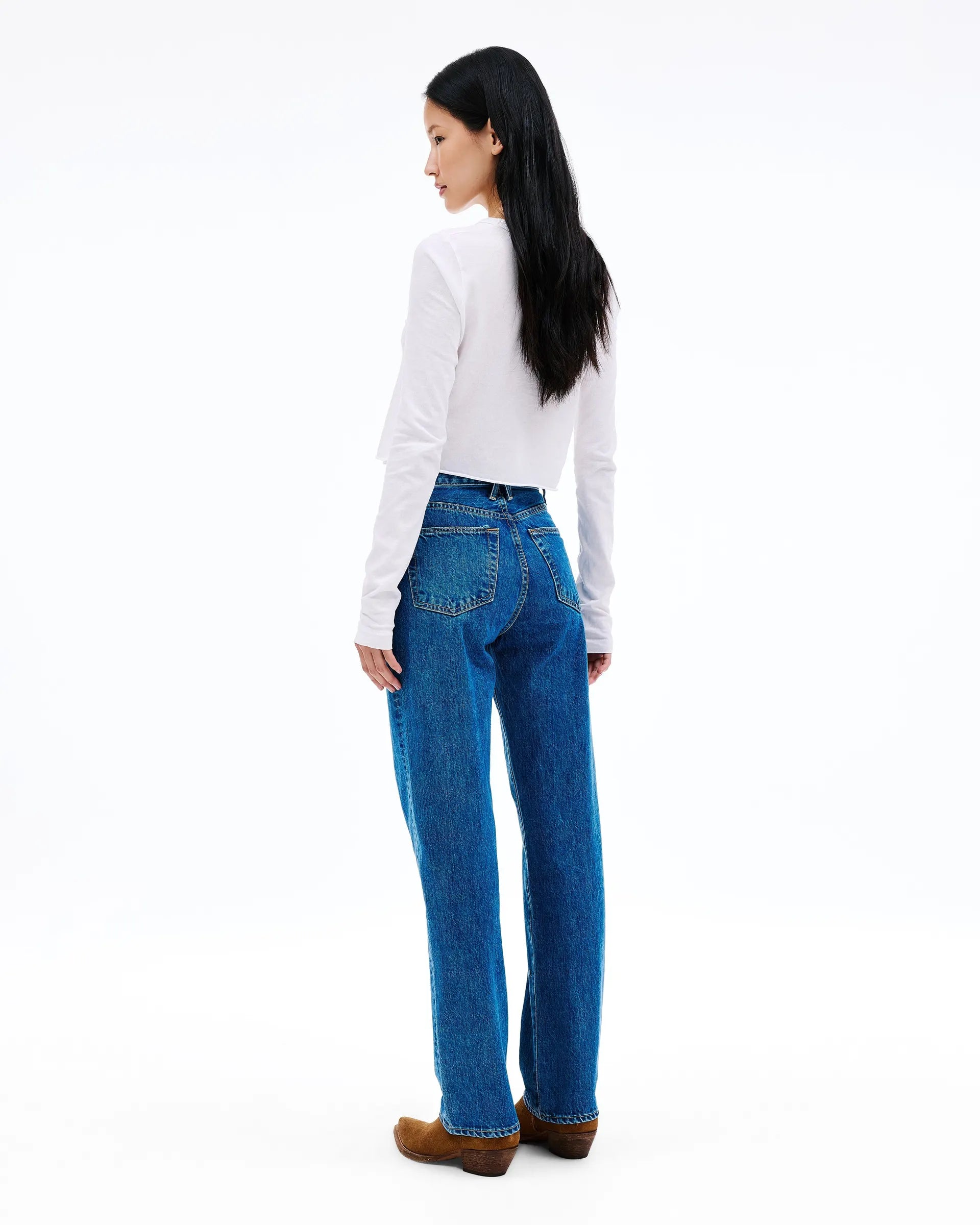 A woman shown from the back wearing a white long-sleeve top, blue high-waisted straight-leg jeans, and brown suede shoes, photographed against a seamless white studio background.