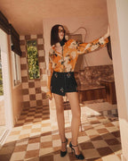 A woman wearing a floral silk blouse with a high collar and long sleeves, standing in a room with a tiled floor and wallpaper.