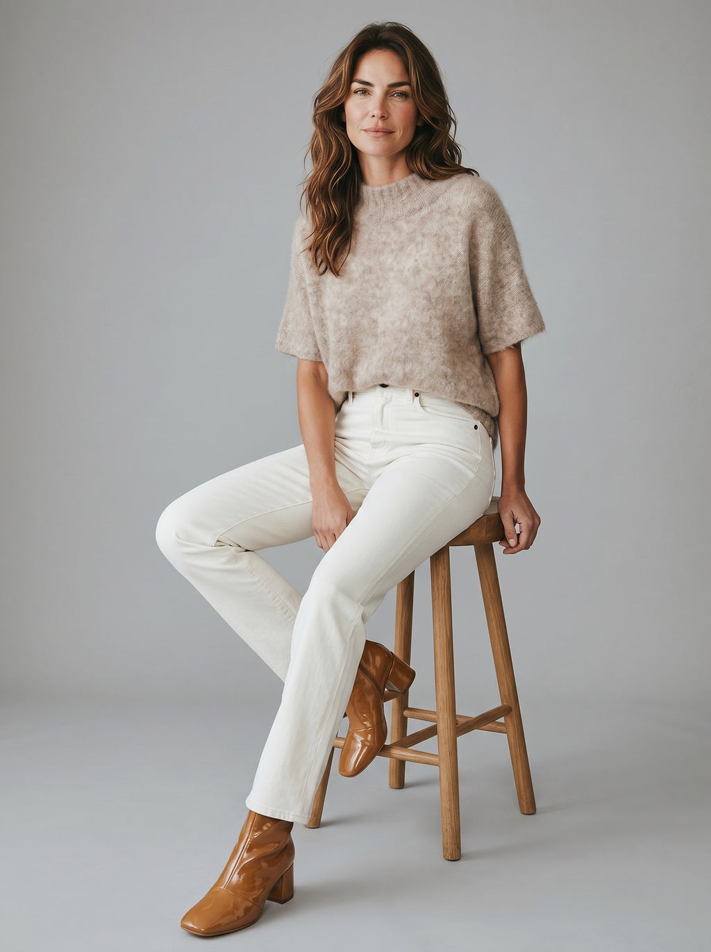 Woman sitting on a stool wearing a beige sweater, white pants, and brown boots against a gray background. rabens saloner. tunic mushroom