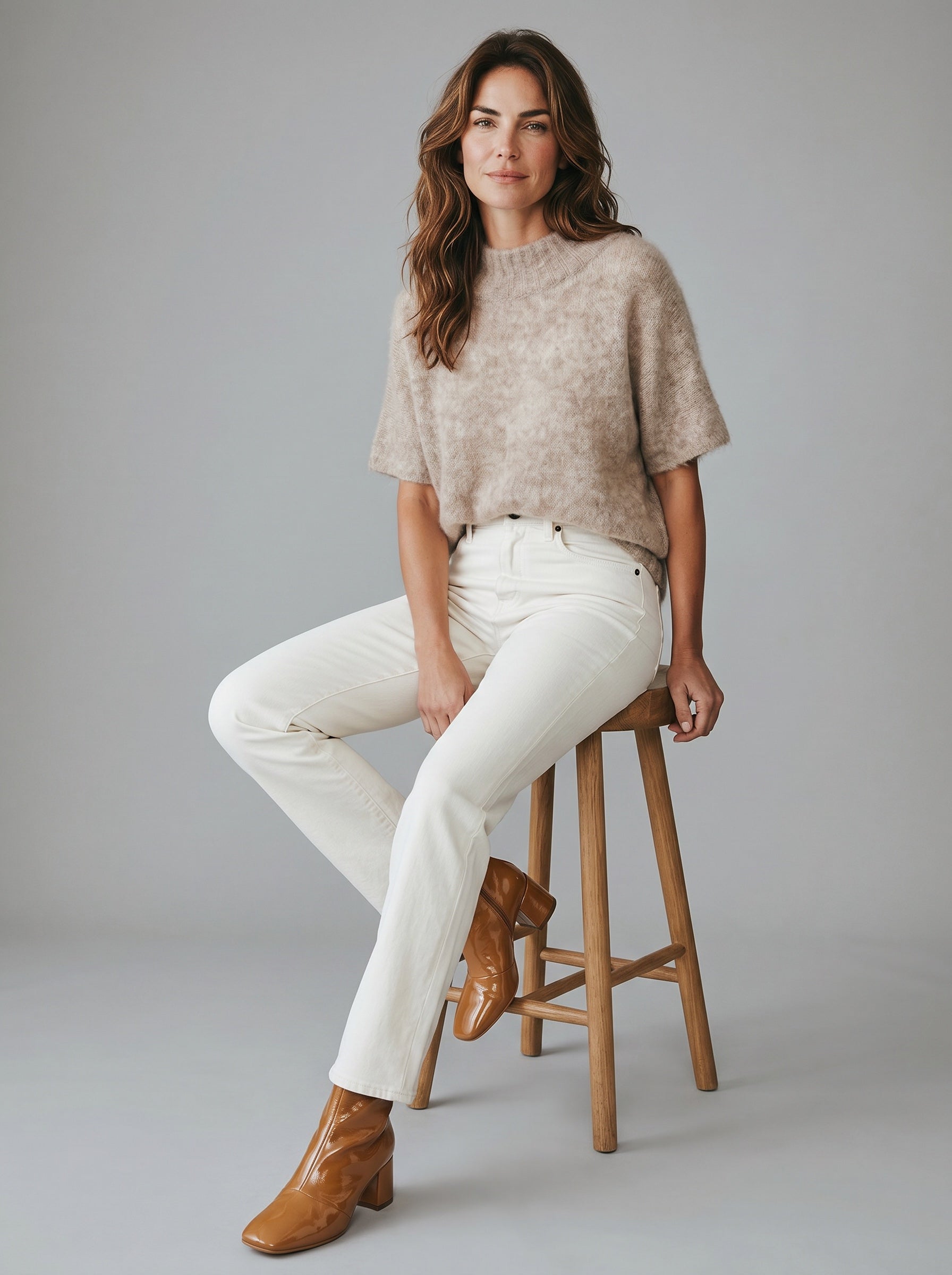 Woman sitting on a stool wearing a beige sweater, white pants, and brown boots against a gray background. rabens saloner. tunic mushroom