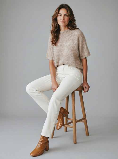 Woman sitting on a stool wearing a beige sweater, white pants, and brown boots against a gray background. rabens saloner. tunic mushroom