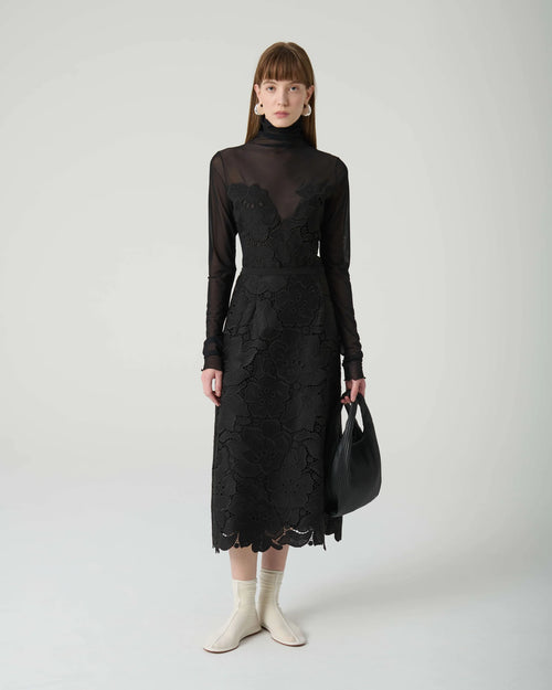 Woman wearing a black lace dress with sheer long-sleeve top against a white background