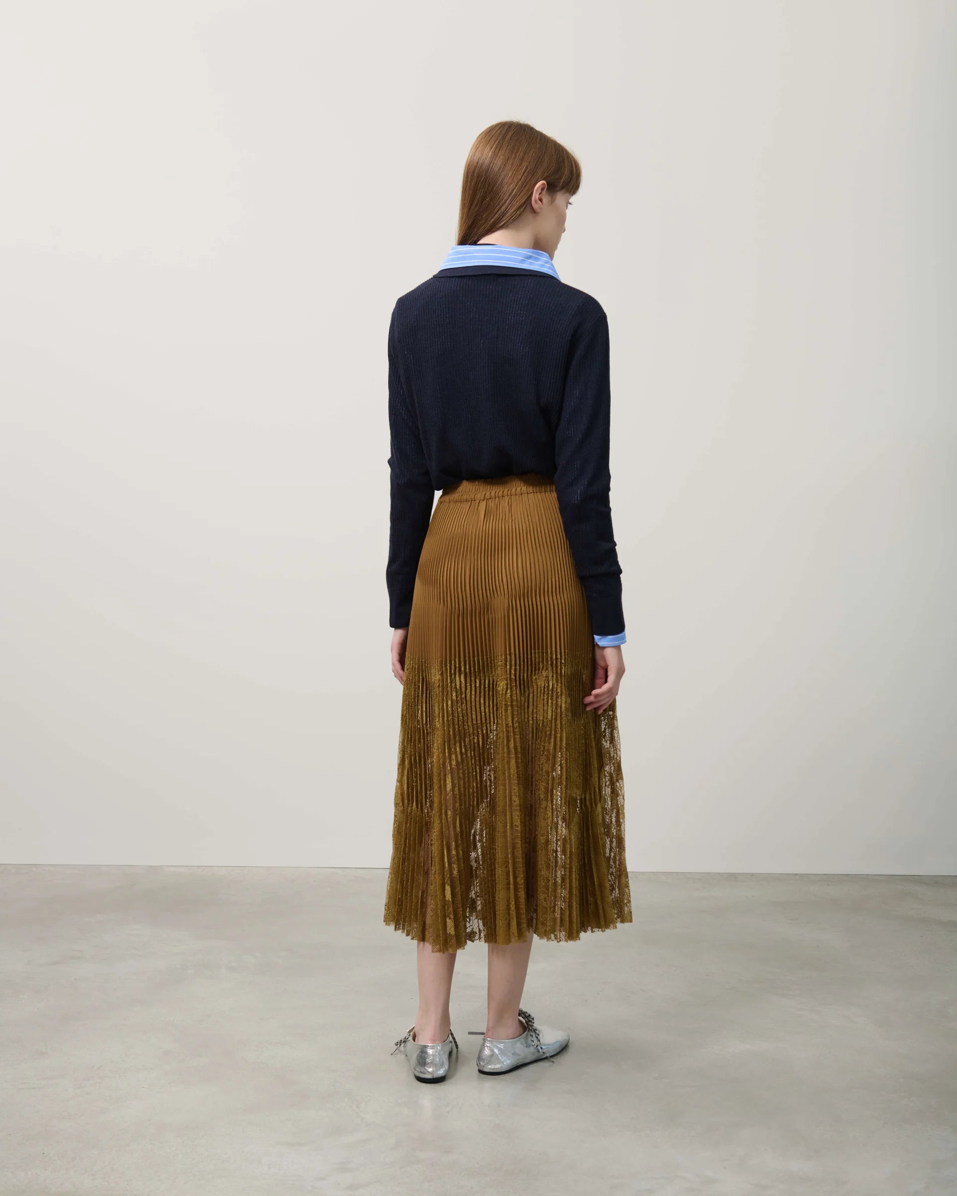 Person wearing a navy sweater and brown pleated skirt against a plain background