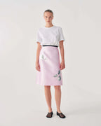 Woman wearing a white t-shirt and pink skirt with floral embroidery on a plain background