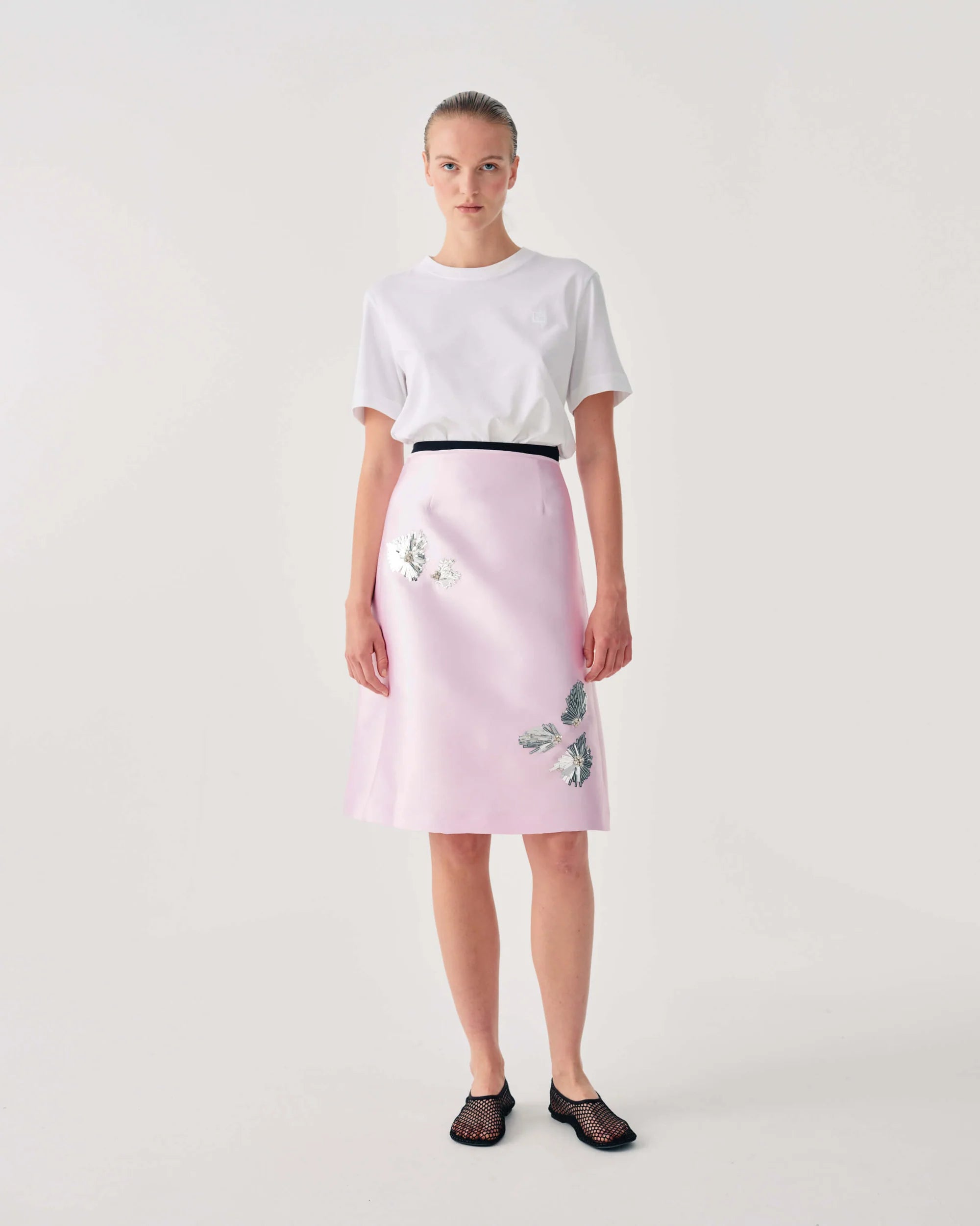 Woman wearing a white t-shirt and pink skirt with floral embroidery on a plain background