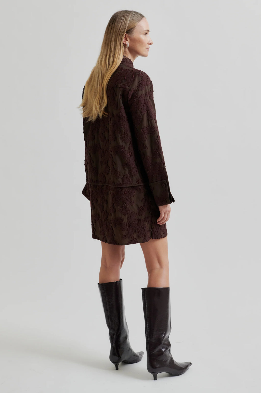 Woman wearing a brown textured outfit with black knee-high boots on a light gray background