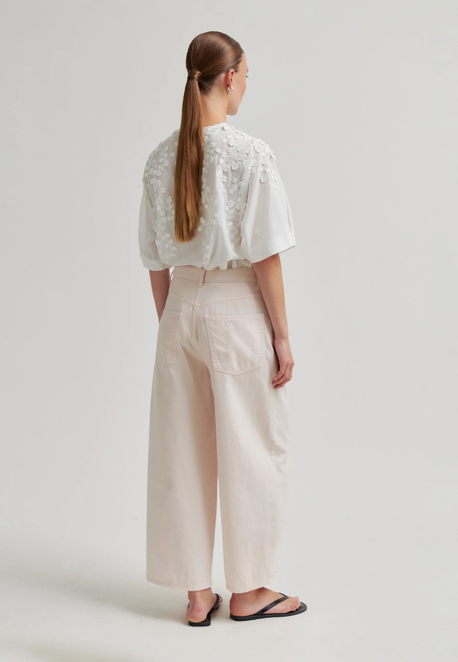 A woman shown from the back wearing a white short-sleeve blouse with floral lace embroidery on the shoulders and high-waisted, wide-leg cream trousers, paired with black flip-flop sandals, against a light gray studio background.