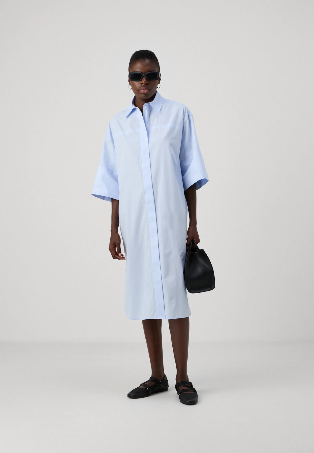 A person standing wearing a Day Birger et Mikkelsen pigeon blue short-sleeved shirt dress with a collar, button closure, and a black handbag.